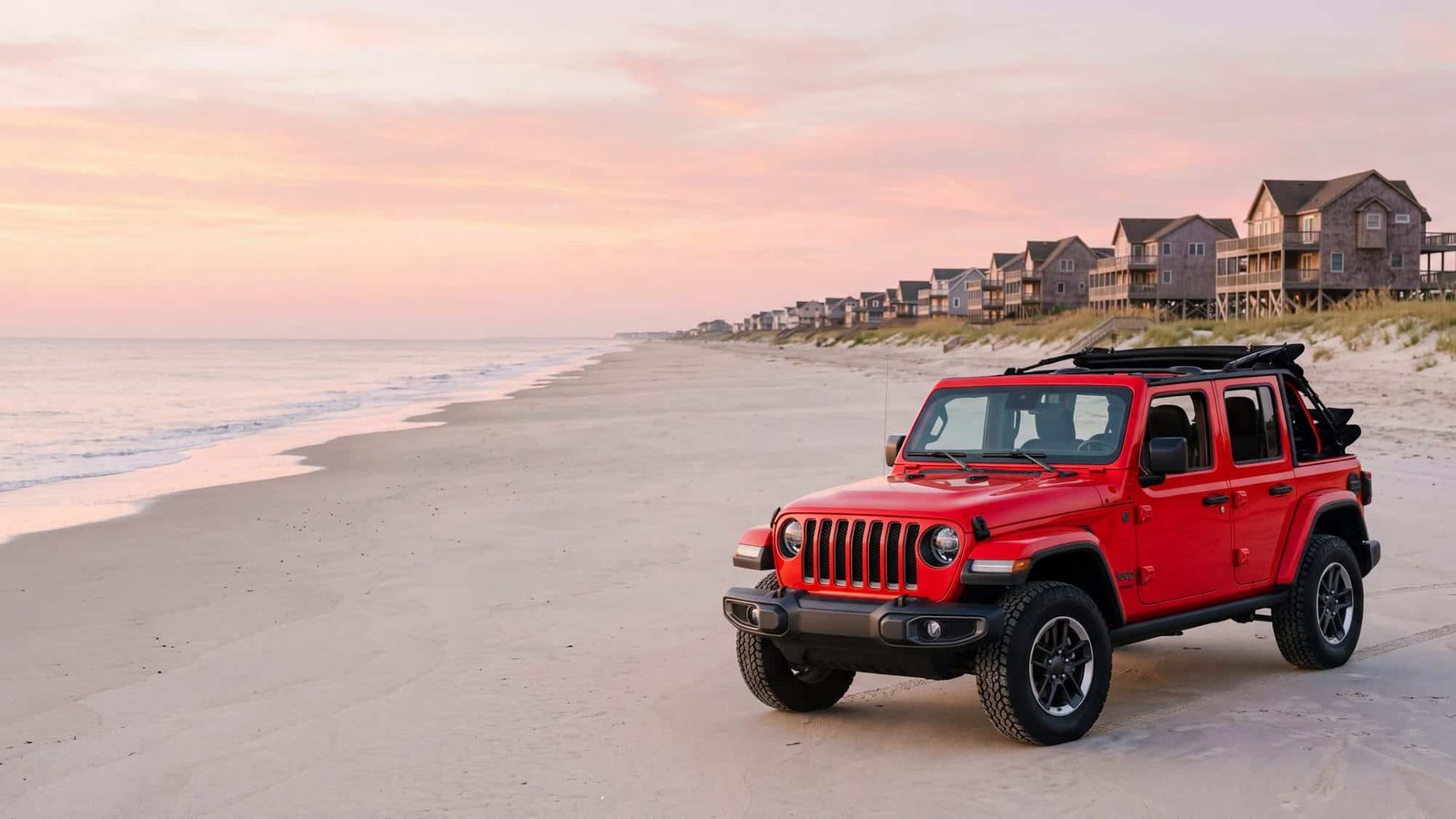 A red Jeep Wrangler parked on the beach at first light with beach houses stretching down the dune line — Carova, Outer Banks NC